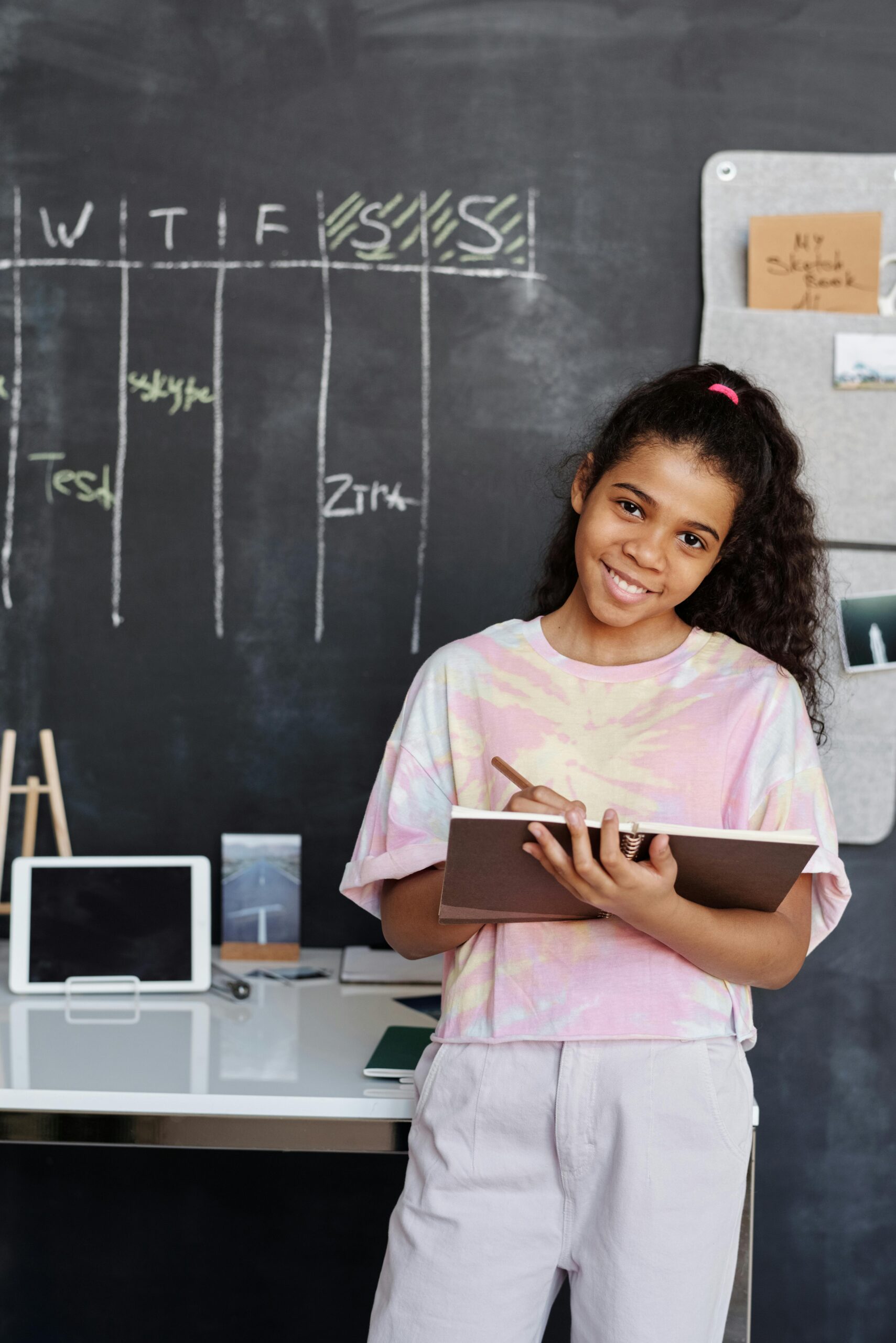 female student looking at camera while holding a notebook in front of a blackboard